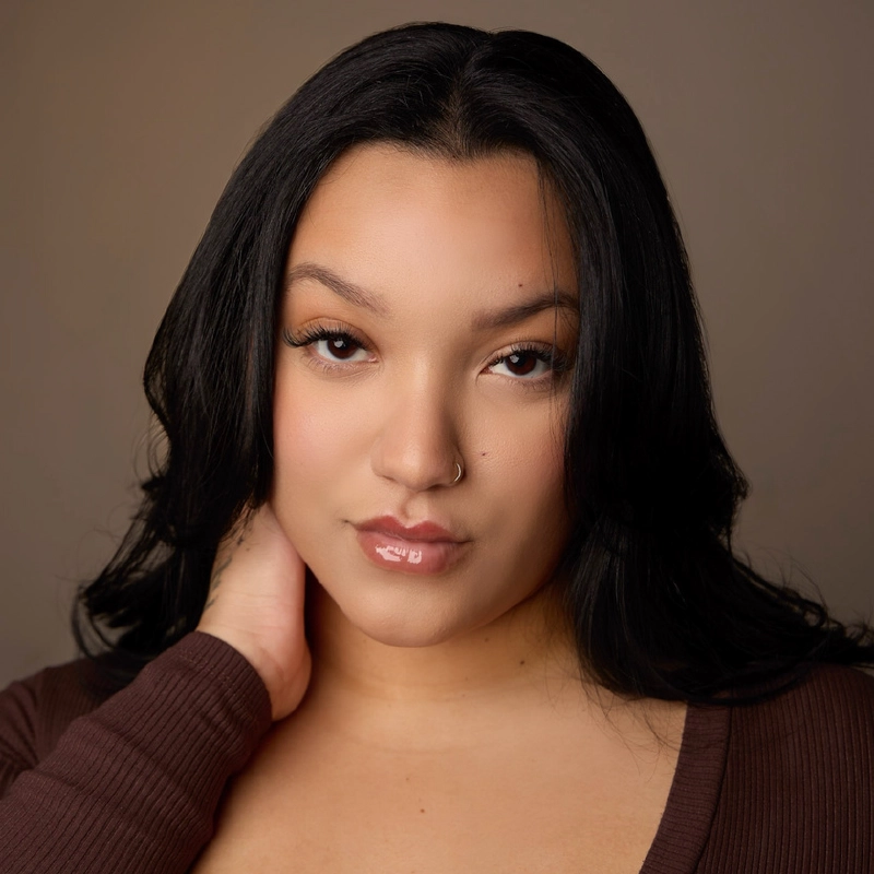A woman with long, dark hair poses against a neutral brown background. She has a subtle, confident expression, light makeup with glossy lips, and a small nose piercing. She wears a brown top and rests one hand gently on her neck.