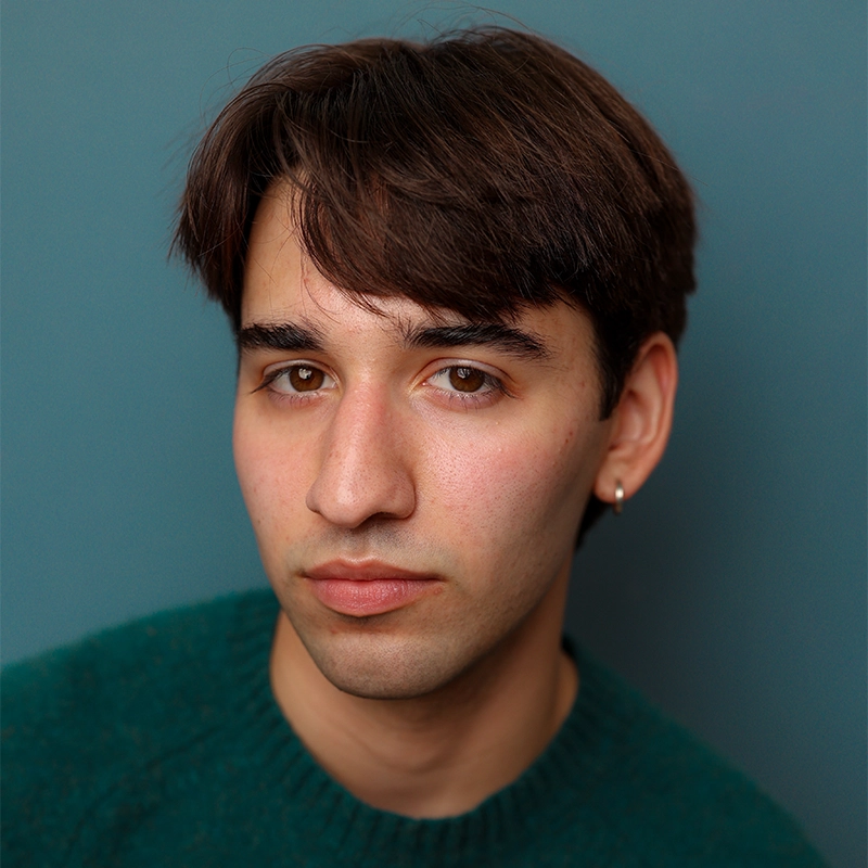 A young person with straight brown hair, light skin, and brown eyes looks seriously at the camera. They wear a green sweater and a small hoop earring in their left ear. The background is a plain blue-gray color.