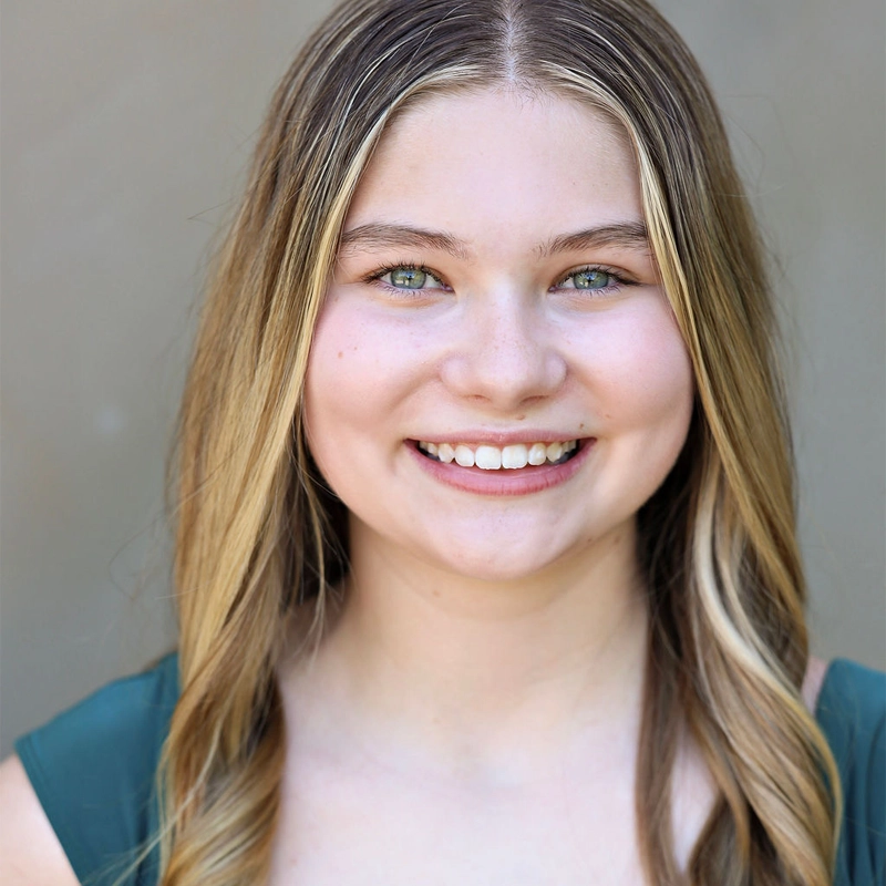 A young woman with long, straight, blonde hair and blue eyes smiles at the camera. She is wearing a green top and standing in front of a neutral, blurred background. The lighting is bright and natural, highlighting her clear skin and cheerful expression.