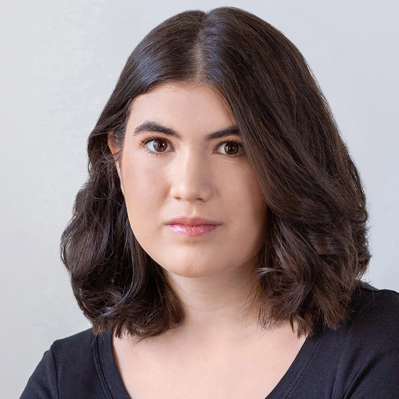 A young woman with medium-length, wavy dark brown hair, brown eyes, and light skin looks directly at the camera with a neutral expression. She is wearing a black top and is posed in front of a plain, light-colored background.