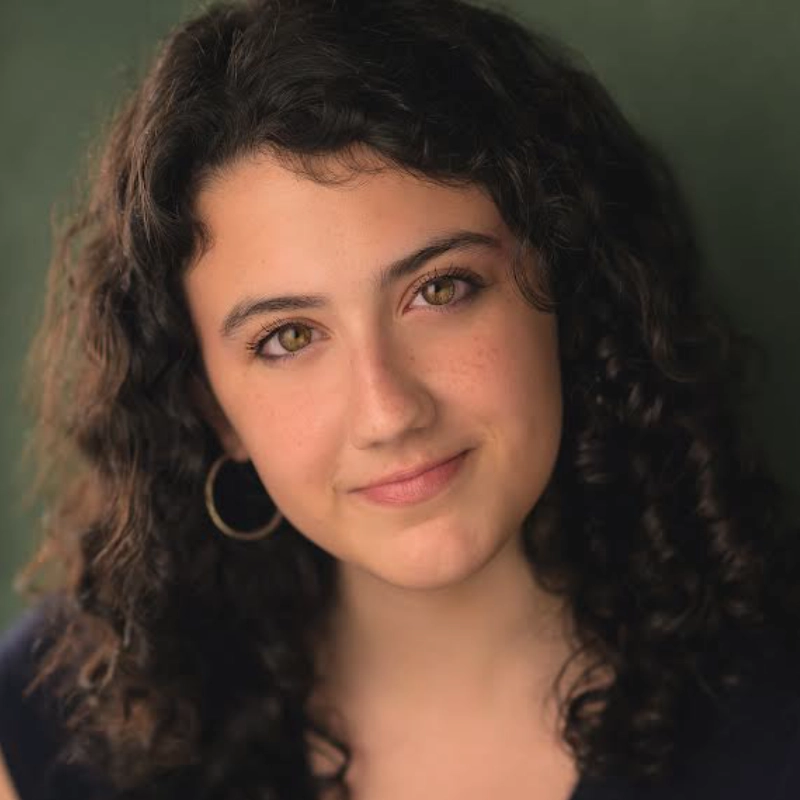 A young woman with curly dark hair, wearing a dark top and a large hoop earring, smiles softly at the camera. She has light skin, hazel eyes, and a faint smattering of freckles. The background is a muted green color.