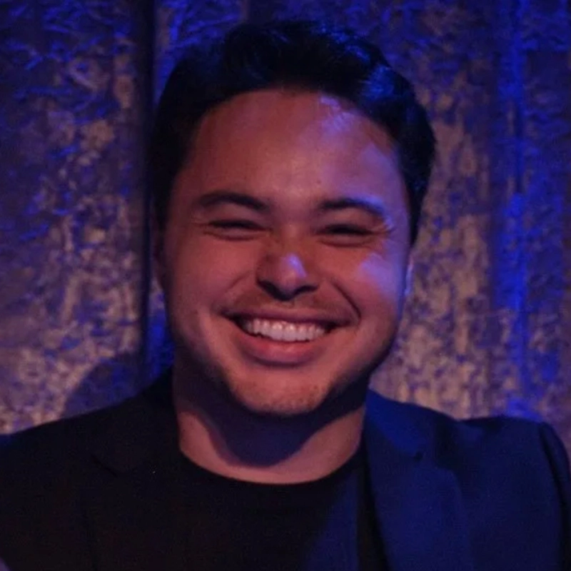 Timothy Fenton, a man with short dark hair, is smiling broadly, showing his teeth. He is wearing a black shirt and jacket. The background is dimly lit with blue lighting and a textured curtain, creating a warm and friendly atmosphere.
