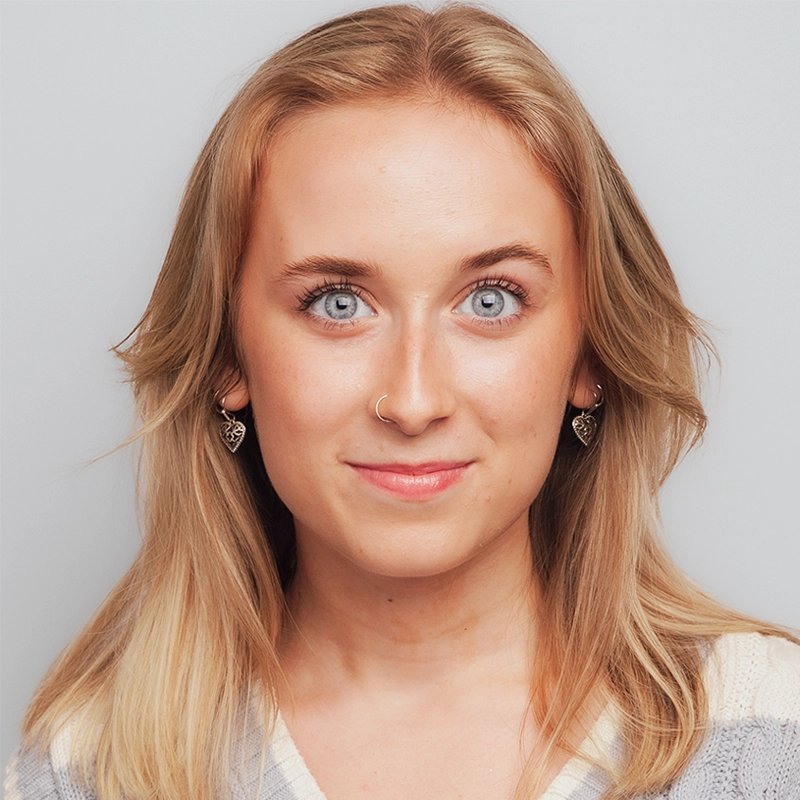 Mary Grace Smith, a young woman with long blonde hair, blue eyes, and fair skin, looks directly at the camera with a slight smile. She has a nose ring, heart-shaped earrings, and wears a light-colored striped top against a light gray background.