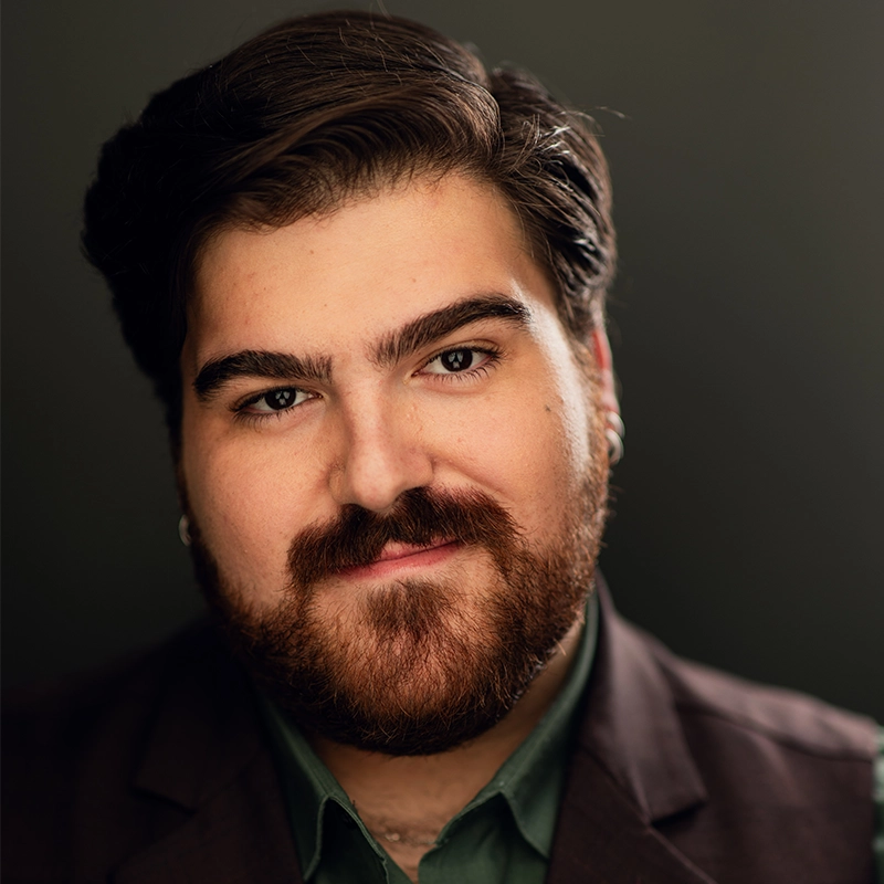 A man with dark, styled hair and a full brown beard smiles slightly. He has thick eyebrows, wears earrings, and is dressed in a dark brown suit jacket over a dark green shirt. The background is dark and softly blurred.