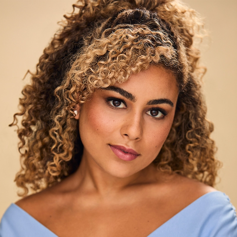 A woman with curly, highlighted hair styled half-up poses against a neutral background. She has medium skin, defined brows, and natural makeup with rosy lips. She wears a light blue off-the-shoulder top and gazes confidently into the camera.