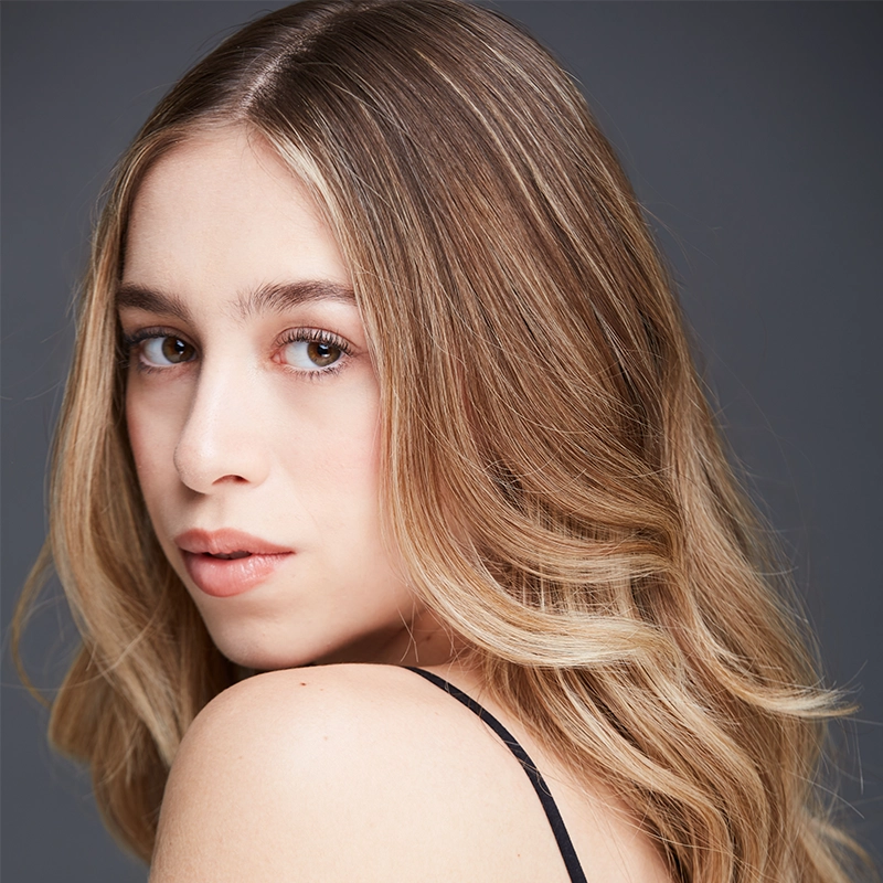A young woman with long, wavy, blonde hair looks over her shoulder toward the camera. She has fair skin, natural makeup, and wears a black spaghetti strap top. The background is a plain, dark grey—her look is reminiscent of Majo Cárdenas.