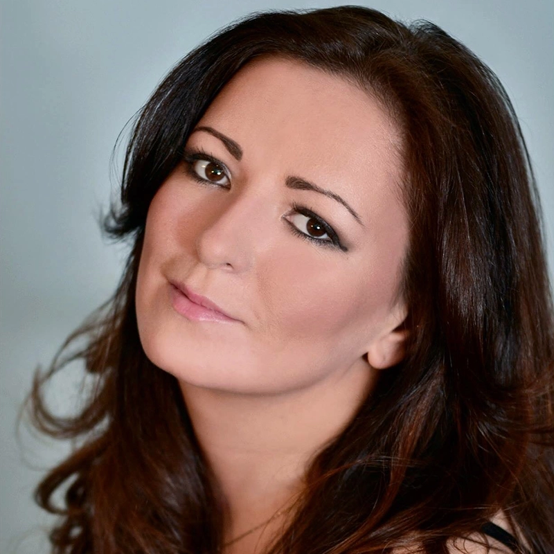 Margaret Kelly, with long dark brown hair and fair skin, gazes softly at the camera. Her brown eyes, subtle makeup, and gentle expression are complemented by a soft blue background, creating a calm and serene atmosphere.