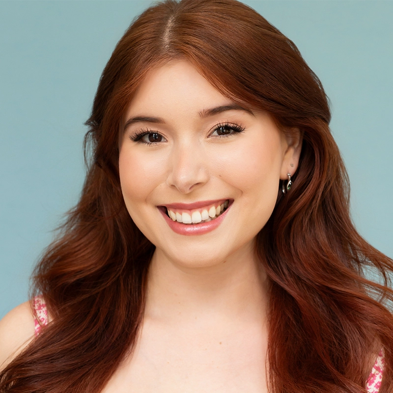 A young woman with long, wavy auburn hair smiles warmly at the camera. She has fair skin, natural makeup, dark lashes, and wears small hoop earrings. Her dress is pink with a subtle pattern, and the background is a solid light blue.