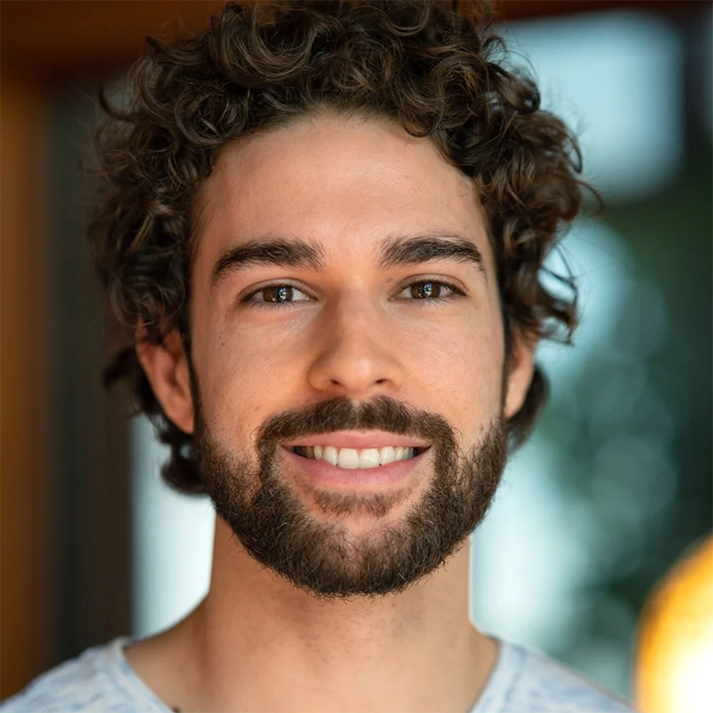 A headshot of a young man with curly brown hair and a full beard smiling at the camera. He has warm brown eyes and is wearing a light-colored, patterned crew-neck shirt against a soft, out-of-focus background.