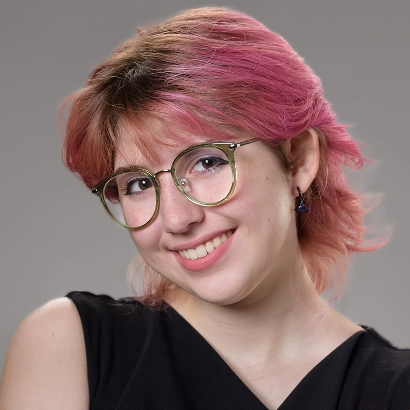 A young person with short, wavy pink hair smiles at the camera. They wear round glasses with light-colored frames, purple earrings, and a sleeveless black top. The background is a neutral gray, and the lighting is soft and even.