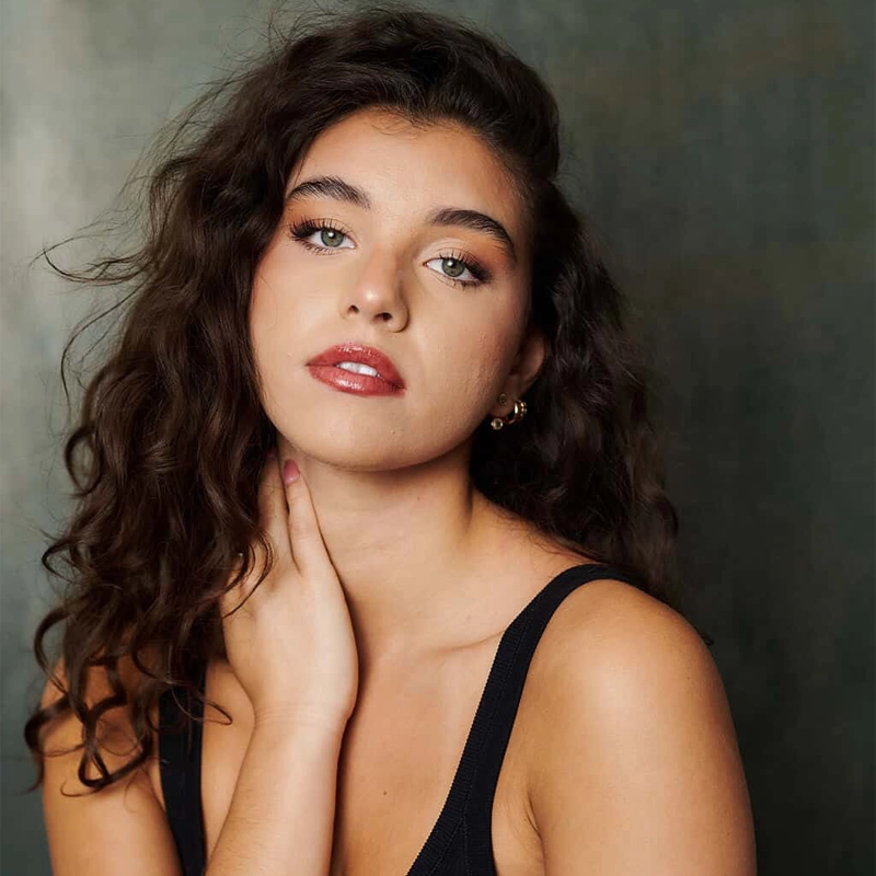 Giovanna Martinez, a young woman with long, wavy brown hair, poses against a muted, dark background. She wears a black sleeveless top, gold hoop earrings, and natural makeup with pink lipstick as she gazes directly at the camera.