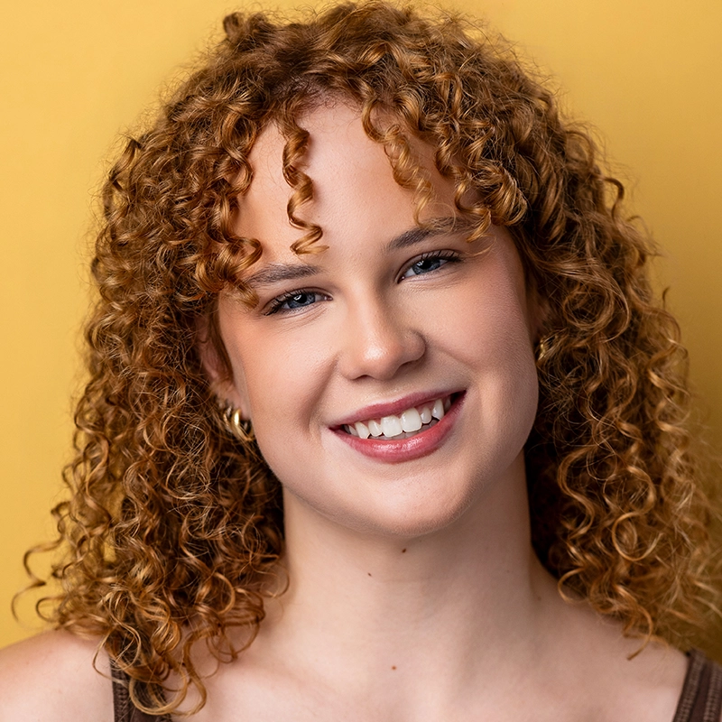 A young woman, Mary Grace Smith, with fair skin and curly, shoulder-length red hair smiles warmly. She has blue eyes, light makeup, and wears small hoop earrings. The solid mustard yellow background creates a bright, cheerful atmosphere.
