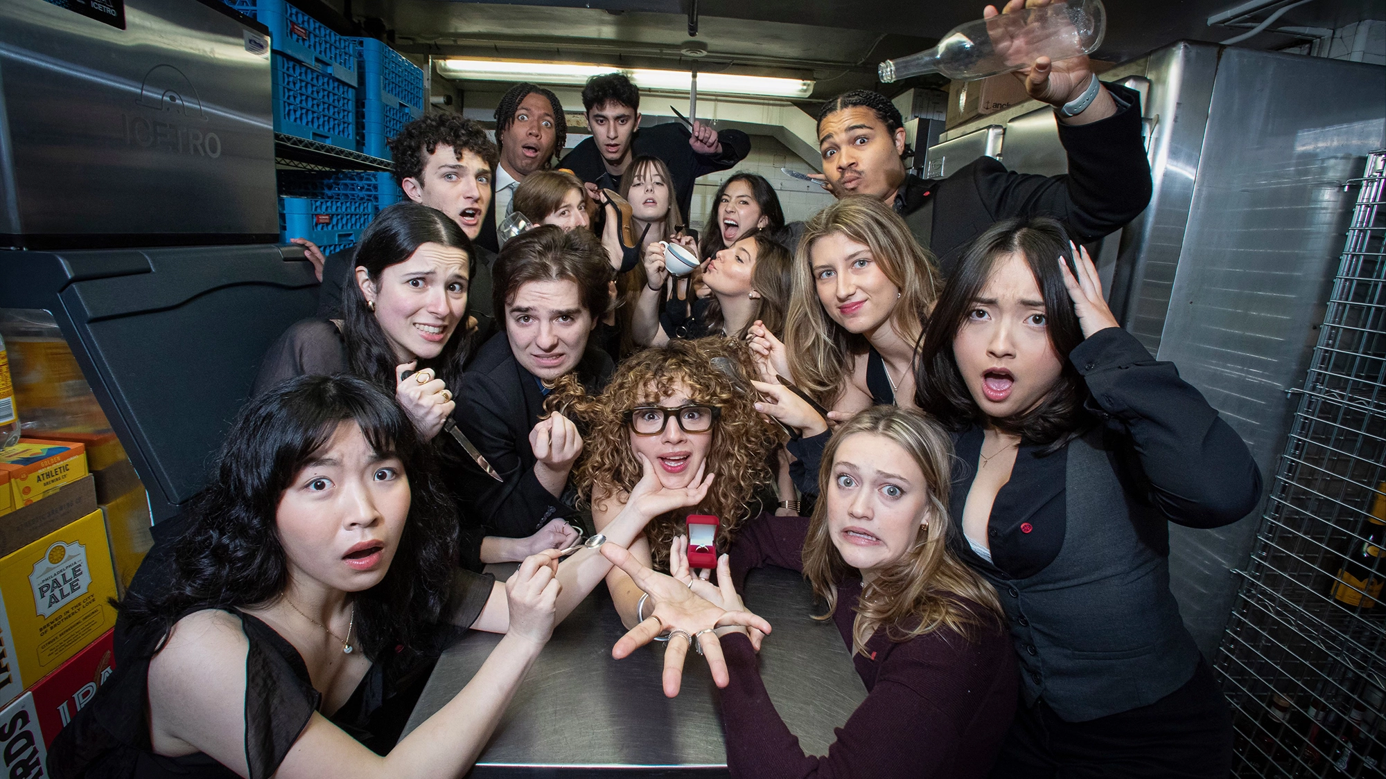 Mask & Wig site image 27 A group of people in black clothing gather in a kitchen, reacting with shock and surprise as one person at the center holds out a ring box. The scene echoes Mask and Wig’s signature flair—everyone gasps dramatically, embodying “Wig You Be Mine” energy.