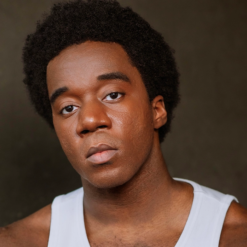 A young Black man with short, curly hair looks seriously at the camera. He is wearing a white tank top, posed against a dark background. His calm, contemplative expression captures the introspective style of photographer Matthew Joseph.