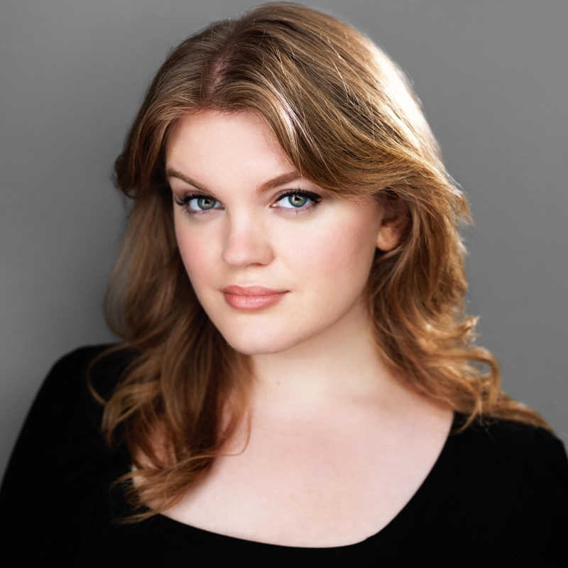 Mia McCarthy, with wavy light brown hair and light eyes, looks at the camera with a soft, confident expression. She wears a black top against a plain gray background, her features highlighted by natural makeup and subtle lighting.