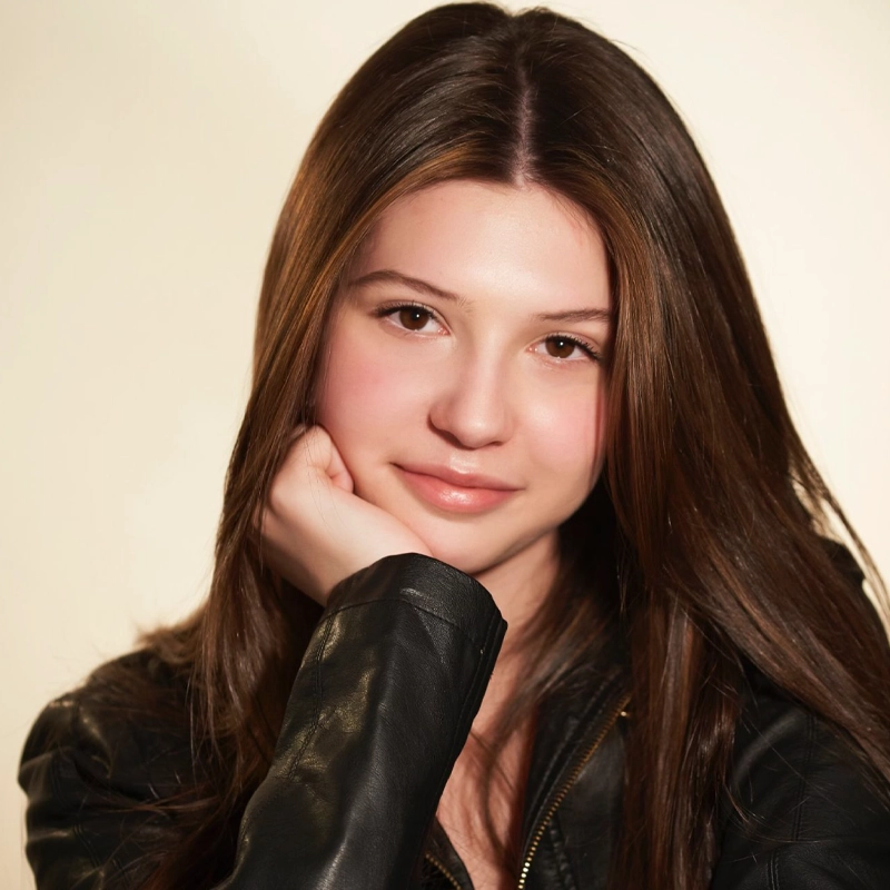 Alana Melillo, a young woman with long brown hair, looks at the camera, resting her chin on her hand. She has fair skin, wears a black leather jacket, and smiles softly against a plain light background.