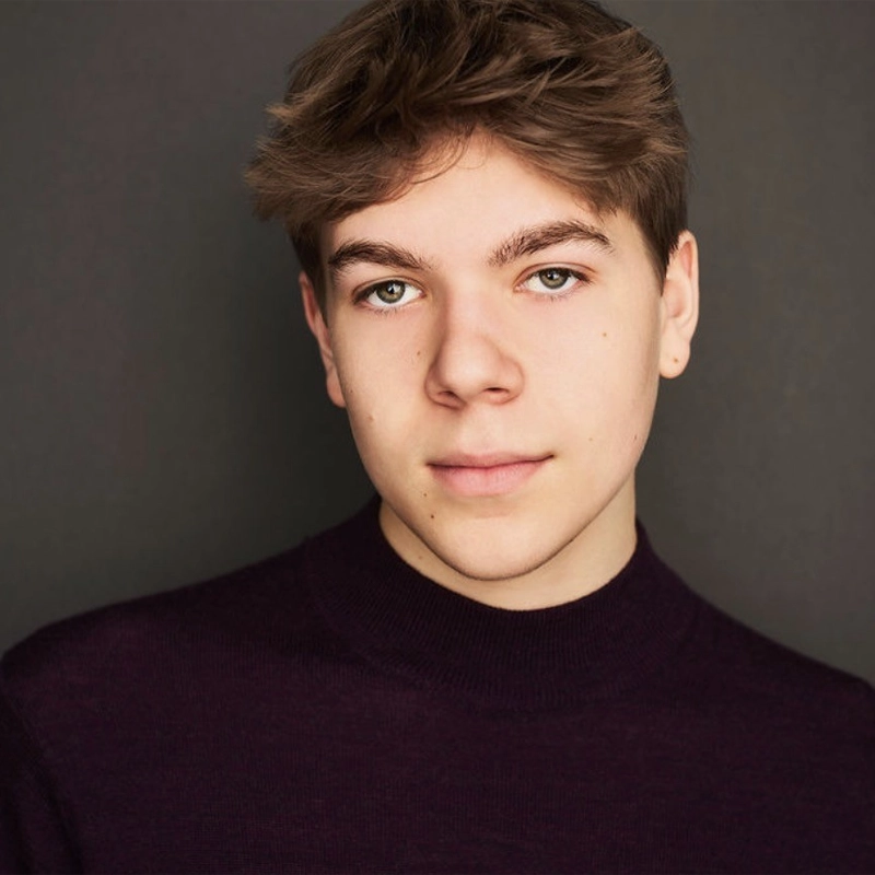 A headshot of a young person with light brown hair and green eyes, looking directly at the camera. They are wearing a dark purple mock-neck sweater against a simple grey background.