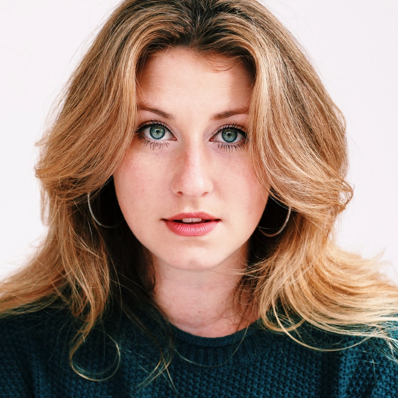 Jessica Morilak, with long wavy blonde hair and light eyes, stares directly at the camera. She wears hoop earrings and a dark textured sweater. The background is a soft white, and her expression is neutral and calm.