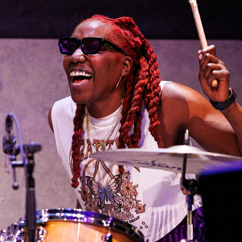 A smiling drummer with long red braids and sunglasses plays the drums energetically. She wears a sleeveless white shirt with a colorful design, layered necklaces, and a wristband, holding a drumstick above the drum kit mid-performance.