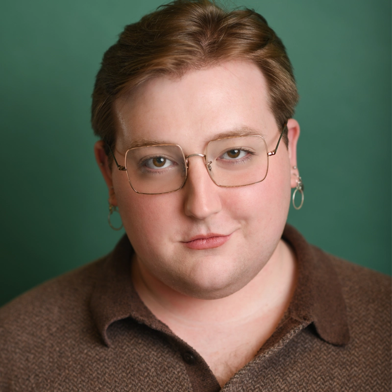 Stephen Nickisch, with short, light brown hair, rectangular glasses, silver hoop earrings, and a brown collared shirt, has fair skin and a neutral expression. The background is a solid green color.