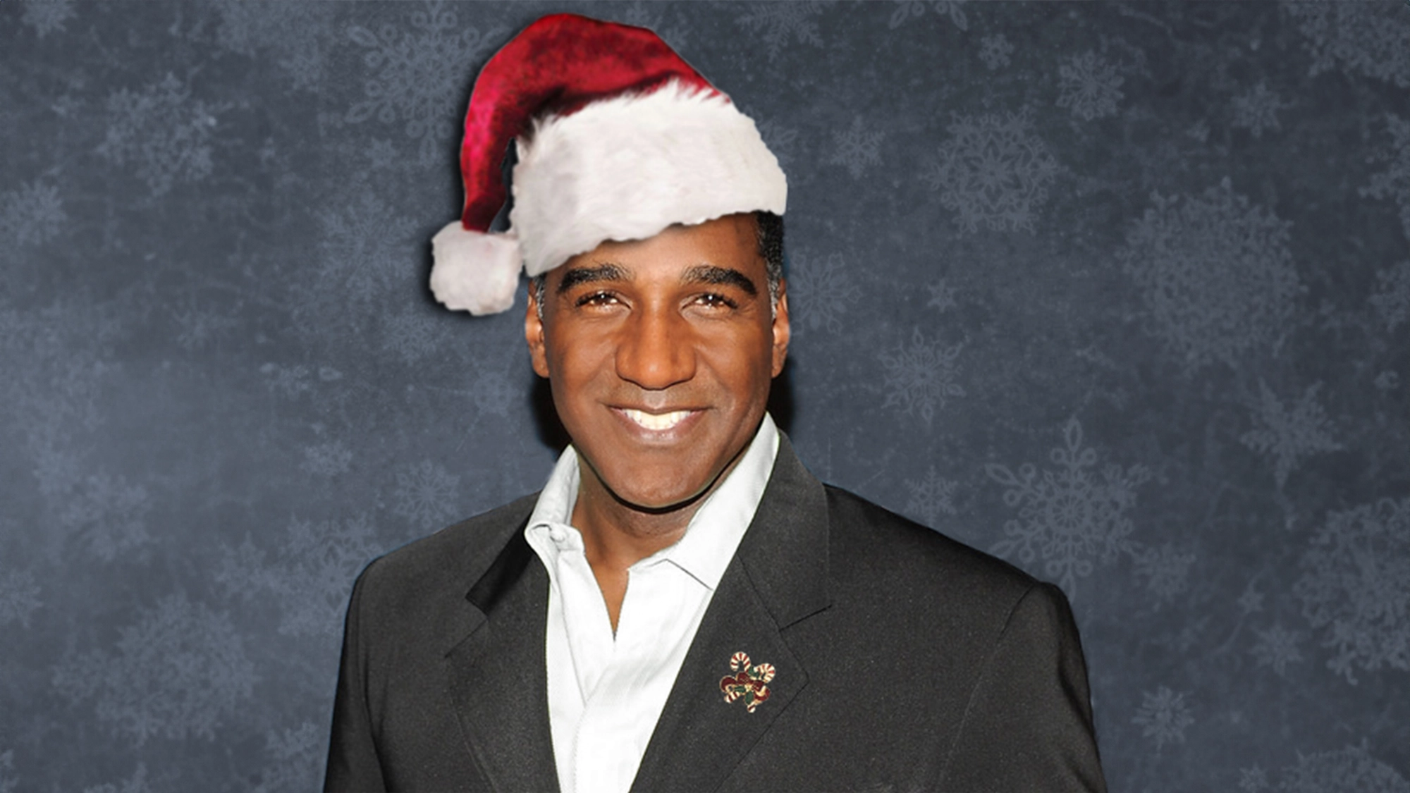 Norm Lewis, wearing a red and white Santa hat and a dark suit jacket with a holiday-themed pin, stands smiling against a textured, dark background with a subtle snowflake pattern—a perfect capture of holiday tradition.