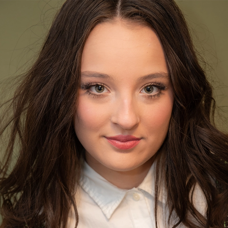 Mary Grace Smith, a young woman with long, wavy brown hair and hazel eyes, looks directly at the camera. She wears subtle makeup and a white collared shirt against a plain, muted green background that draws focus to her face.