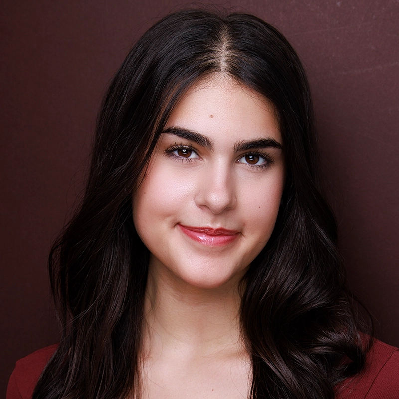 A young woman with long, dark brown hair and fair skin is smiling gently at the camera. She has well-groomed brows, natural makeup, and glossy lips. The background is a smooth, deep burgundy color.