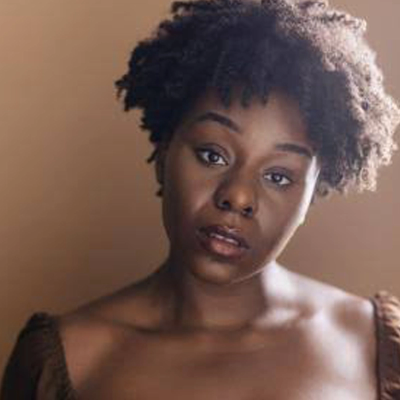 A young woman with short, natural curly hair looks directly at the camera against a soft brown background. Reminiscent of Abigail Onwunali, she wears a brown, off-the-shoulder top and has a calm, thoughtful expression with natural makeup and warm lighting.