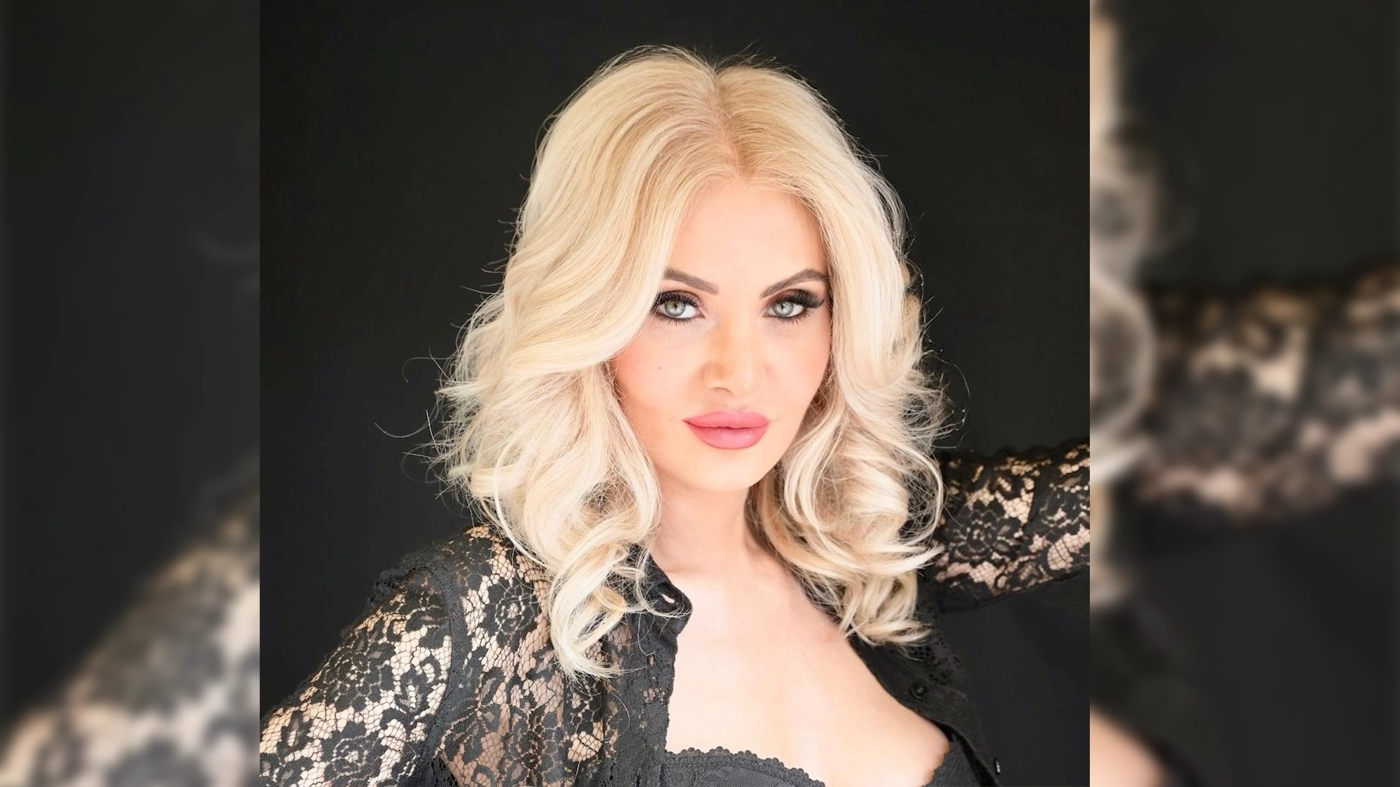 A woman with long, wavy platinum blonde hair poses unbound against a black background. She has light skin, defined eyebrows, dramatic eye makeup, and pink lipstick. Wearing a black lace top with a scalloped neckline, she gazes directly at the camera.
