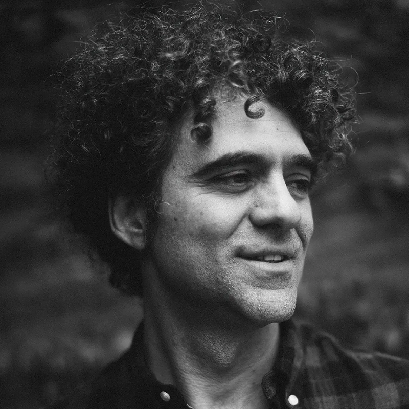 Black and white close-up photo of Peter Salett, a man with curly hair wearing a plaid shirt, looking slightly to his left with a soft smile. The blurred background draws focus to his face, creating a calm and contemplative mood.