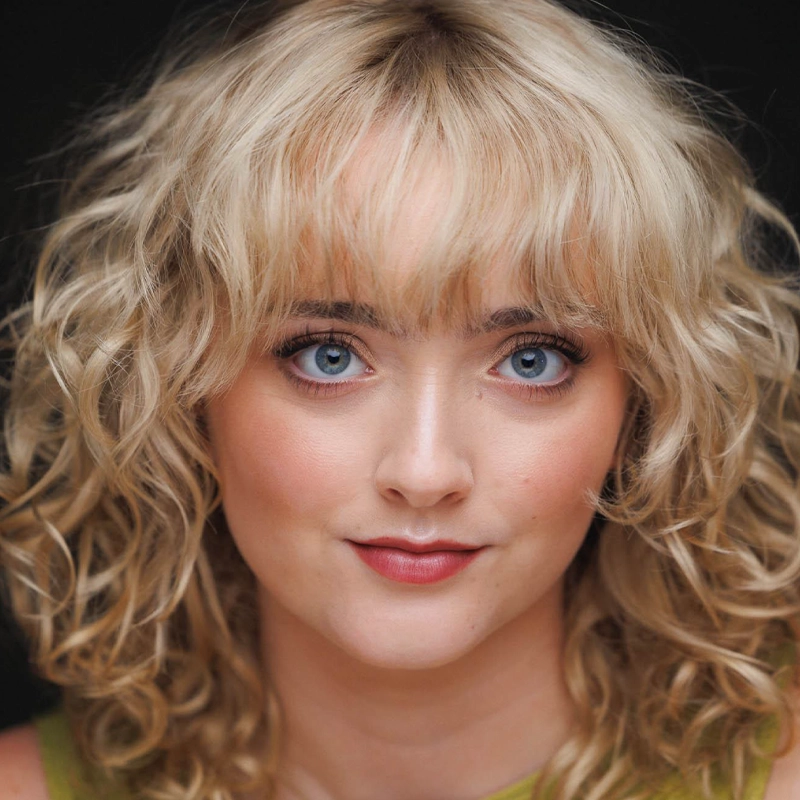 A young woman with light skin and curly blonde hair with bangs stares forward. She has blue eyes, subtle eye makeup, and rosy lipstick. She wears a sleeveless green top, and the background is dark, making her features stand out.