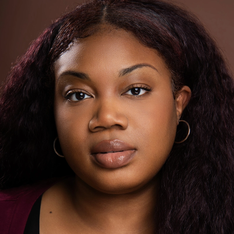 A young woman, Abenaa Quiin, with long, wavy dark hair and medium-brown skin looks directly at the camera. She wears hoop earrings and a dark top, her neutral expression set against a plain brown background.