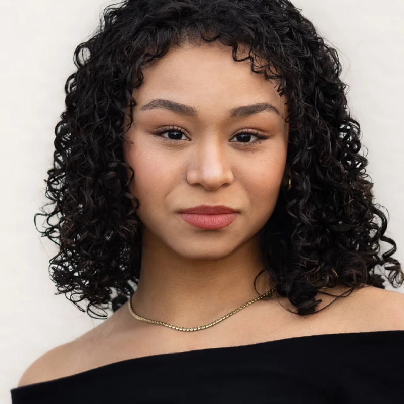 Fabiola Caraballo Quijada, a young woman with curly dark hair, wears a black off-the-shoulder top and thin gold necklace, looking confidently at the camera against a light background. Her natural makeup complements her calm, composed expression.