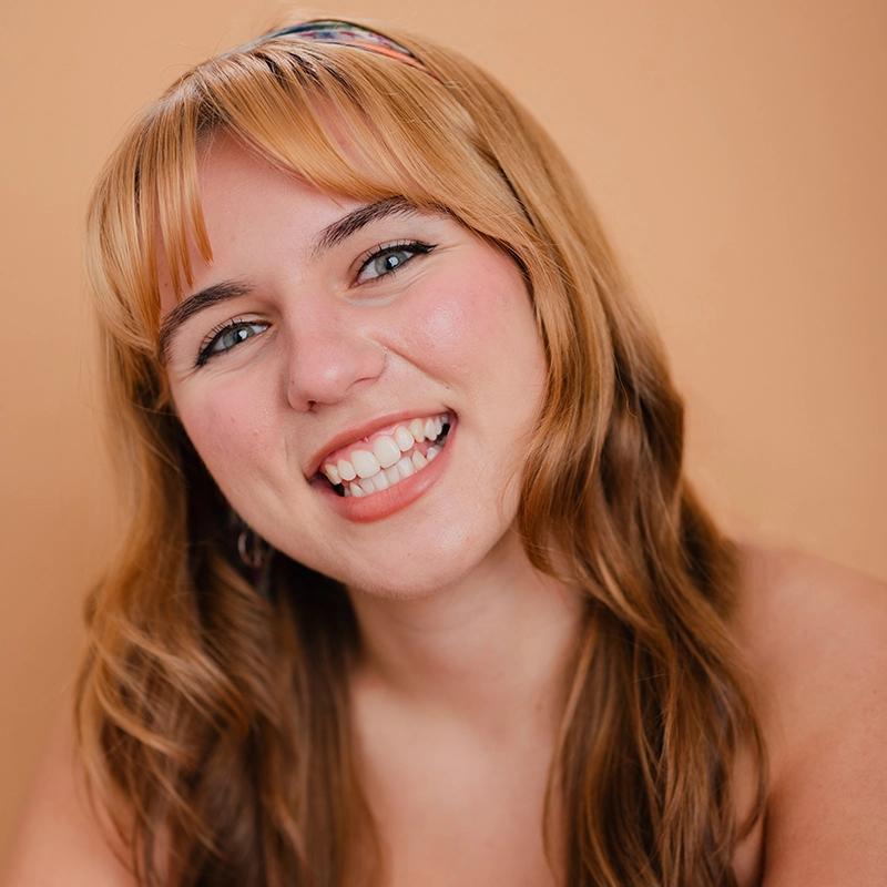 A young woman with long, wavy blond hair and bangs smiles widely, showing her teeth. She is wearing a colorful headband, natural makeup, and is positioned against a plain peach-colored background. Her expression is cheerful and friendly.