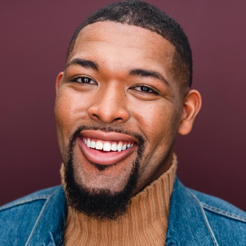 A smiling man with a close-cropped haircut and goatee wears a tan turtleneck sweater and blue denim jacket. He is facing the camera against a solid maroon background. The lighting highlights his cheerful expression and white teeth.