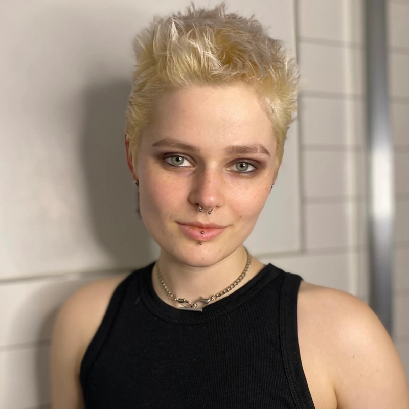 A person with short, spiky bleached blonde hair wears a black sleeveless top and layered chain necklaces. They have piercings on their lower lip and nose, subtle makeup, and are standing indoors against a tiled wall, smiling slightly.