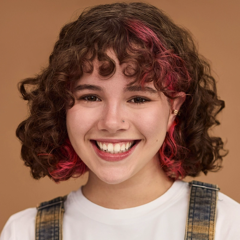 A young woman with curly brown hair featuring pink highlights smiles warmly. She has a nose stud, wears a white shirt with denim overalls, and stands against a plain beige background. Her expression is cheerful and inviting.