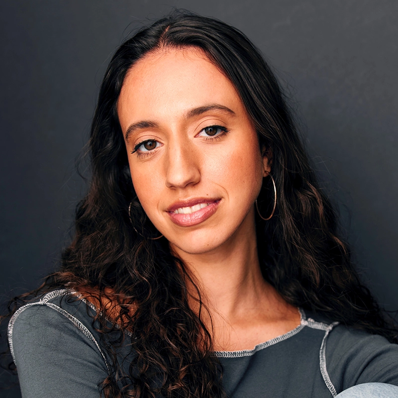 A young woman with long, dark, wavy hair and hoop earrings smiles softly. She wears a gray top with exposed seams and poses against a dark neutral background, giving a confident and relaxed expression.