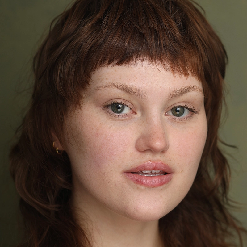 A young person with fair skin, light green eyes, and medium-length, tousled brown hair with short, choppy bangs. They have a natural look with minimal makeup, pink lips, and wear a small gold hoop earring. The background is a muted olive green.