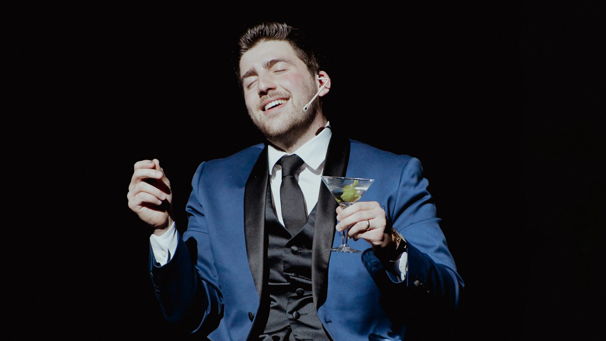 A man in a blue tuxedo with a black lapel and black tie, reminiscent of the American Songbook era, smiles with his eyes closed. He holds a martini glass in one hand and gestures with the other, embodying Ryan Fogler’s signature journey onstage.