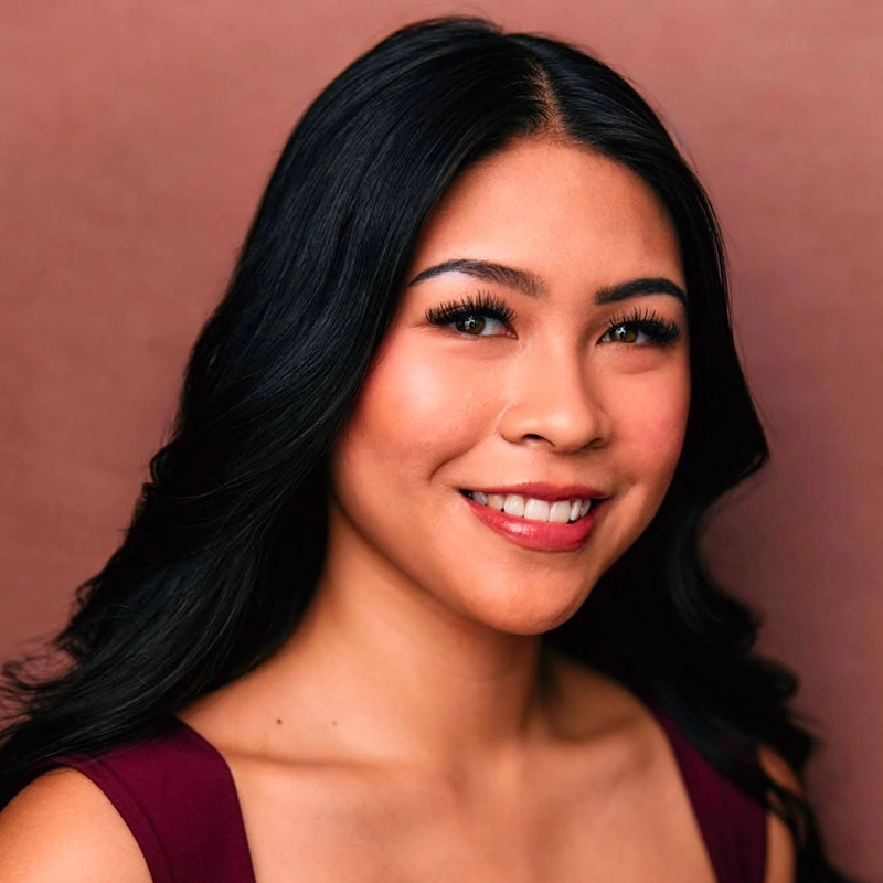 A young woman with long, wavy black hair smiles at the camera. She wears natural makeup with defined brows and lashes, and a burgundy top. The warm brown background creates a gentle, professional portrait—perfect for Cortlandt Barrett’s portfolio.