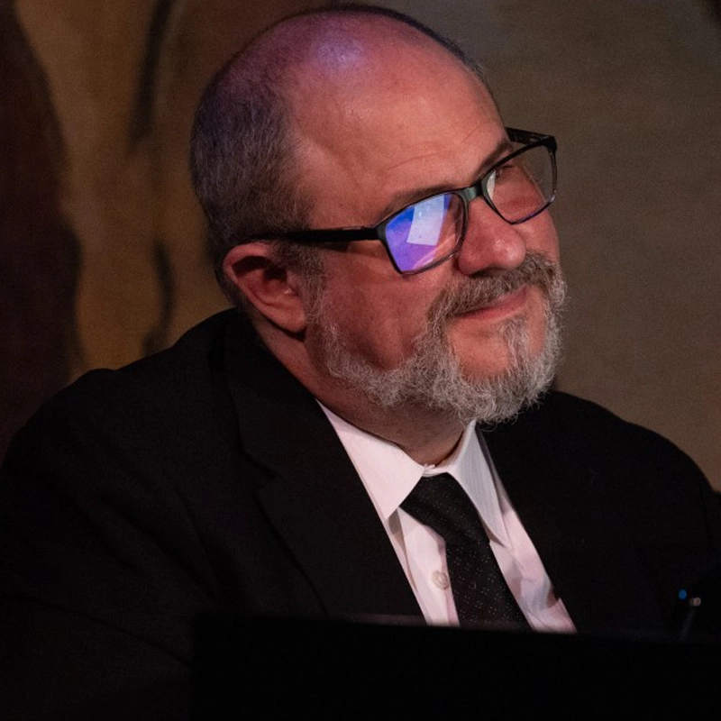 A middle-aged man with a bald head, gray beard, and glasses—James Sampliner—is wearing a black suit, white shirt, and black tie. He is smiling softly and looking off to the side. Warm lighting highlights his face against a dark, blurred background.