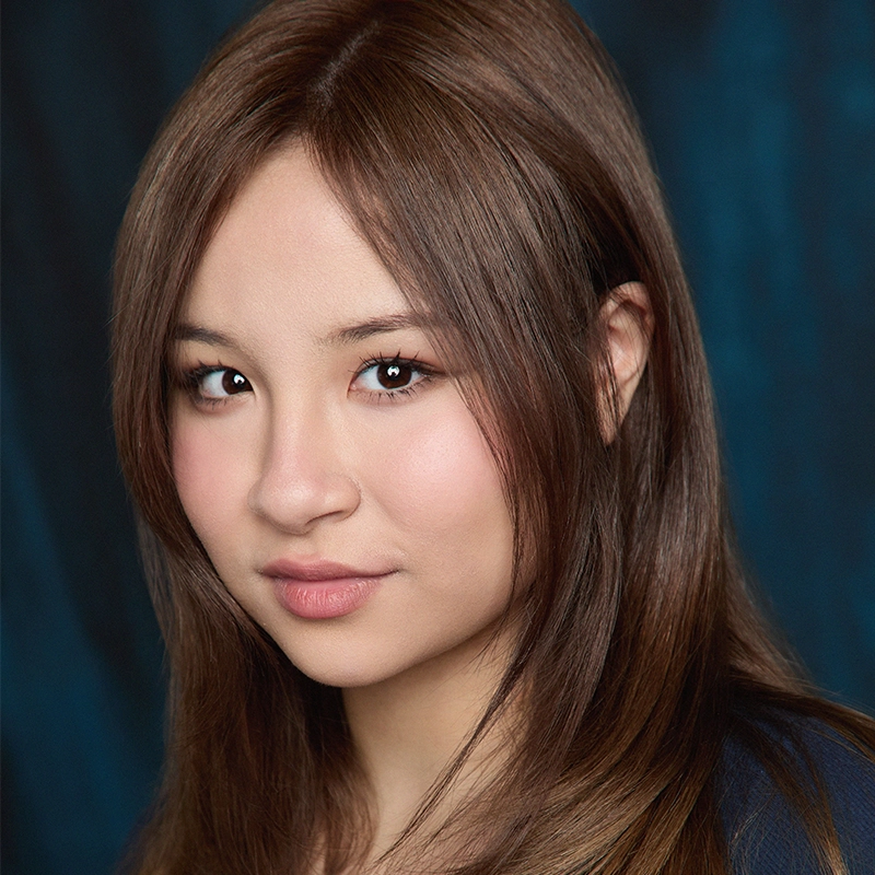 Mary Grace Smith, a young woman with straight, light brown hair and fair skin, gazes at the camera with a soft smile. She has brown eyes, natural makeup, and wears a dark top. The blurred dark blue background highlights her face.