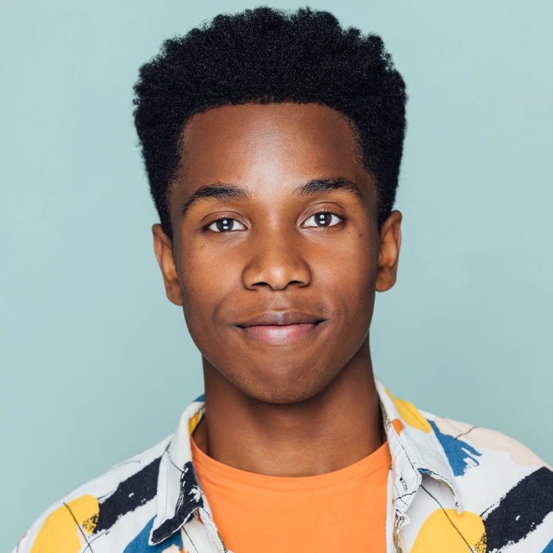 Joshua Screen, a young Black man with short, curly hair, smiles softly. He wears a multicolored, abstract-patterned shirt over an orange t-shirt. The solid light blue background and bright lighting highlight his face.