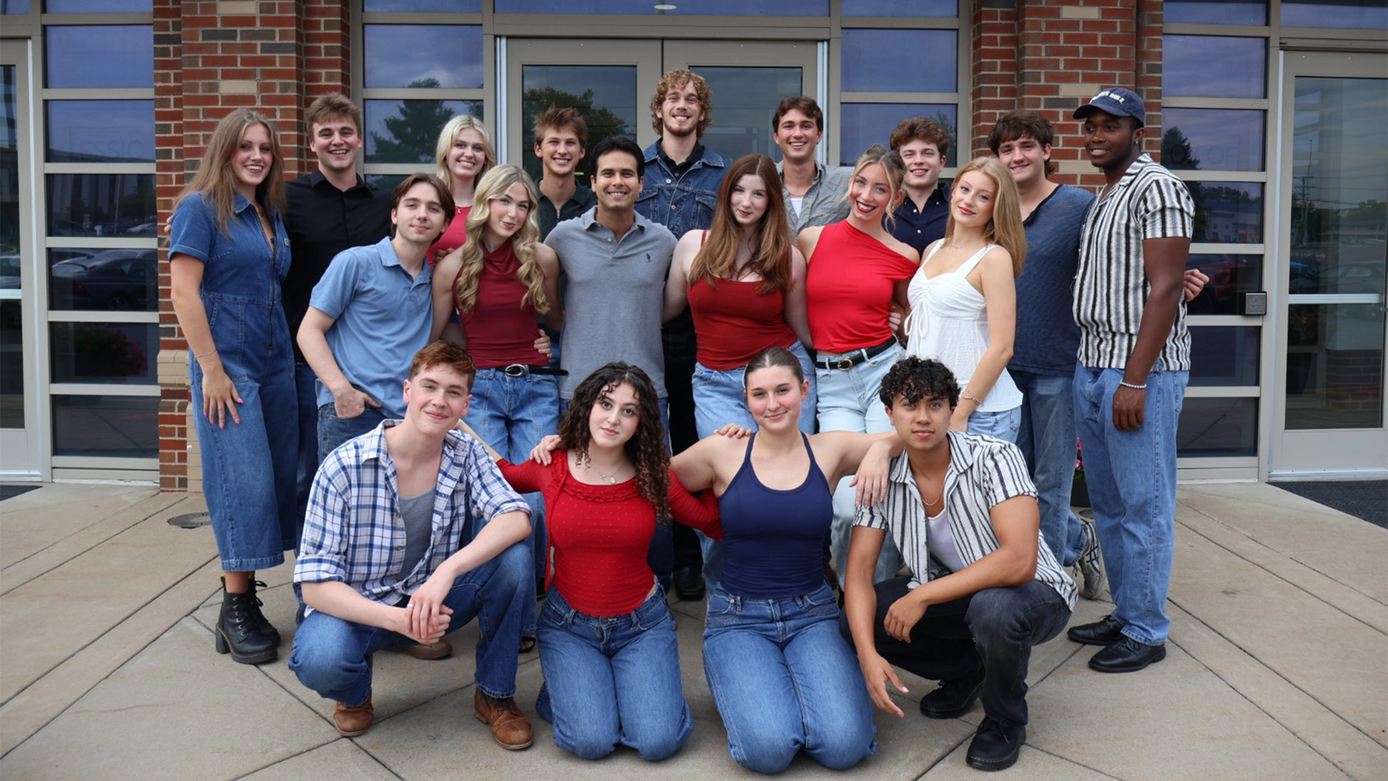 Shenandoah site image 27 The BFA Musical Theatre Class of 2029 poses in front of a brick building with glass double doors. Four people sit in the front row, while the rest stand behind them, all wearing casual shirts and denim. They are smiling at the camera on a sunny day outdoors.