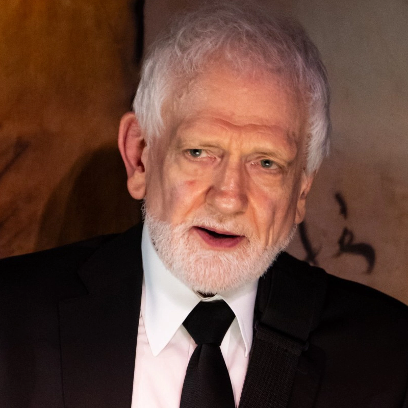 An older man with white hair and a beard wears a black suit, white shirt, and black tie. He appears to be speaking or singing, with a guitar strap over his shoulder. The background is softly lit in brown and beige tones.