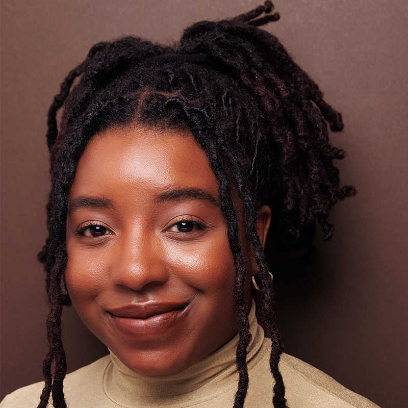 A woman with medium-brown skin and long, thick black locs styled in a high ponytail smiles gently. Sia Michal Arvinger wears a beige turtleneck sweater and gold hoop earring, with natural makeup, against a brown background.