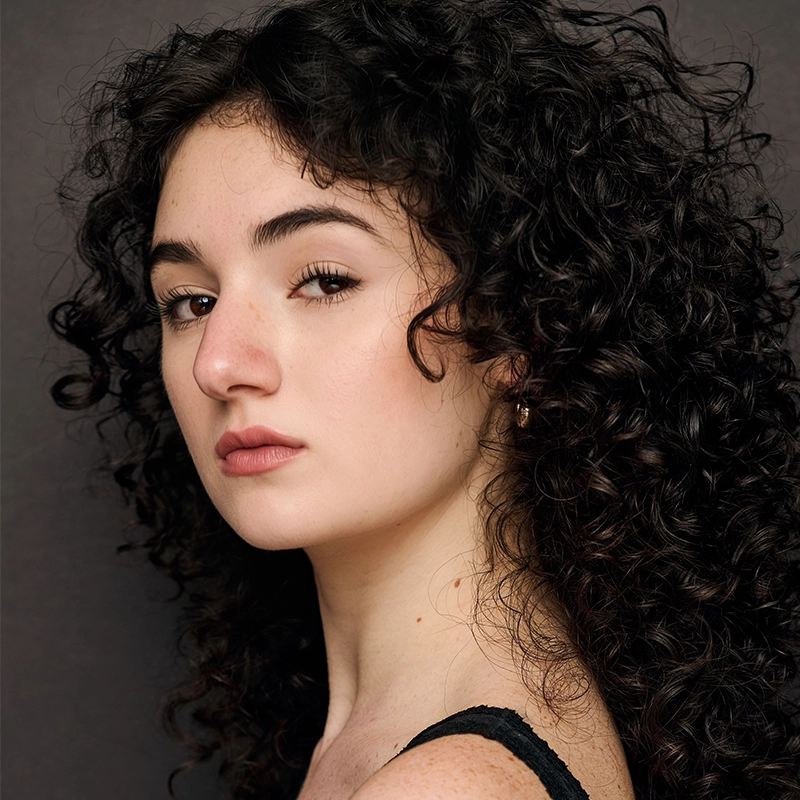 A young woman with pale skin and dark, curly hair looks over her shoulder toward the camera. She has dark eyebrows, brown eyes, and a neutral expression. She is wearing a black top with thin straps, and the background is dark and plain.