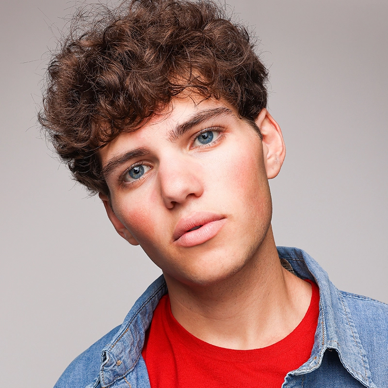 Skyler Reed Lipkind, a young person with curly brown hair and blue eyes, looks into the camera with a neutral expression. They wear a red shirt under a blue denim jacket against a plain, light gray background.