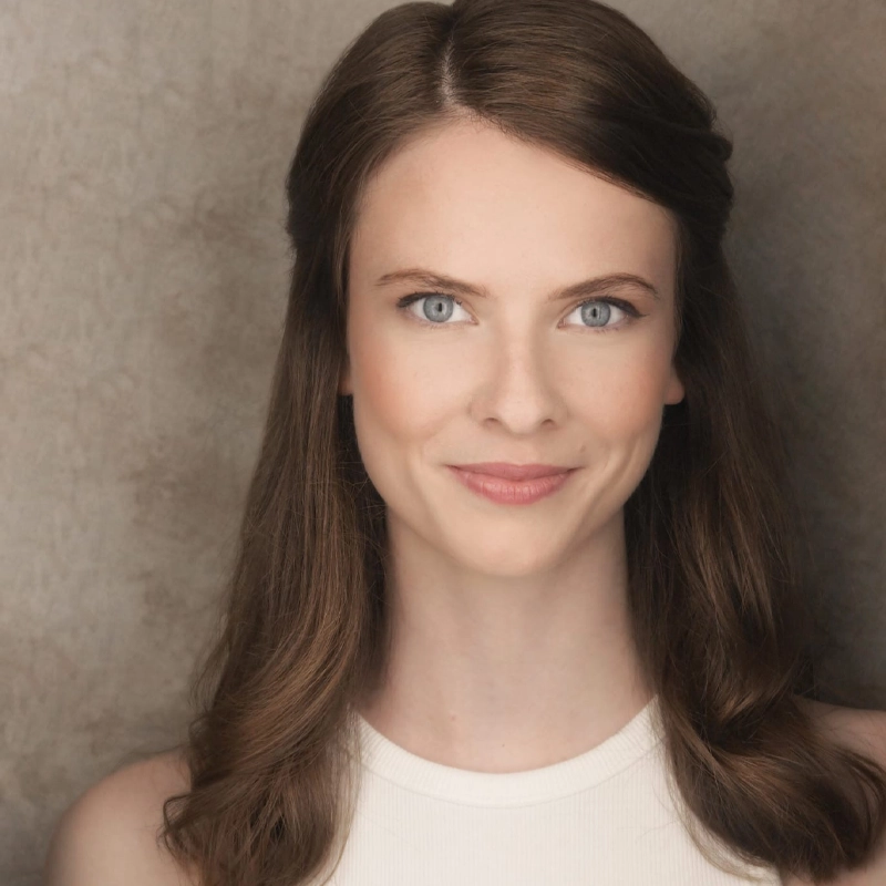 Valentine Smith-Vaniz, a woman with fair skin and long brown hair styled in soft waves, wears a sleeveless white top and smiles softly at the camera. Her blue eyes stand out against the neutral background, enhanced by soft, even lighting.